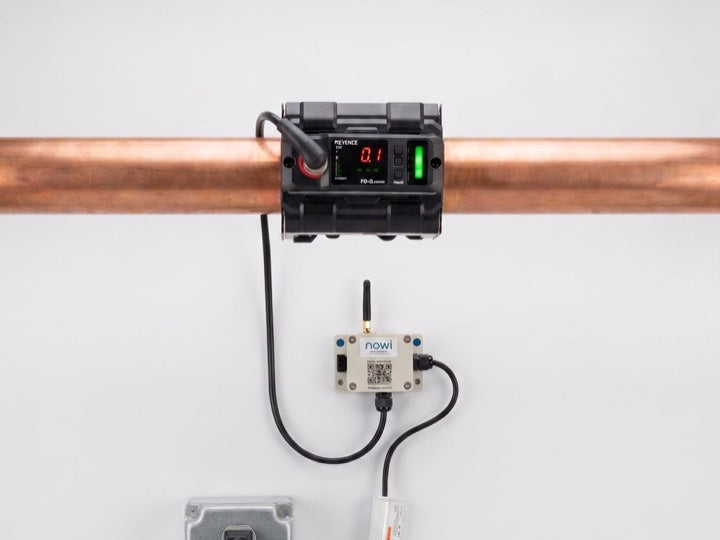 A clamp-on flow sensor on a copper pipe, wired to a NOWi monitor and a wall outlet